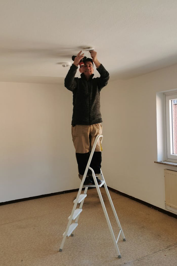 services-06 Adult man on ladder fixing a ceiling light in an empty room with beige walls.
